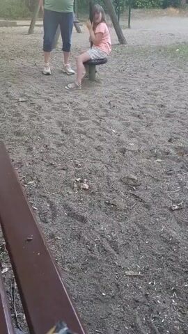 A young girl with dark hair, wearing a pink t-shirt and white shorts, sits on a swing. Her hands are clasped together in front of her face. Her legs are bent at the knees, and her feet are flat on the ground. Behind her, to the left, an adult, possibly a woman, stands with her legs apart, wearing a green t-shirt and blue pants. The ground is covered in sand and small pebbles. In the foreground, a portion of a brown wooden bench is visible, with a crumpled bag of potato chips resting on it. The background shows a swing set and some trees. The overall scene appears to be a park or playground.