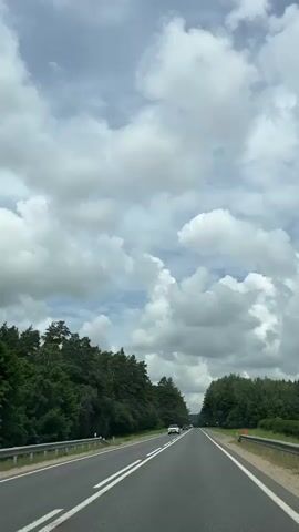 The video shows a first-person perspective of driving on a highway. The road is paved with white lines marking the lanes. On either side of the road are dense green forests with tall trees. The sky is filled with fluffy white clouds against a blue background. Several cars are visible on the road, moving in the same direction as the camera. The overall scene depicts a scenic drive through a natural landscape on a cloudy day.