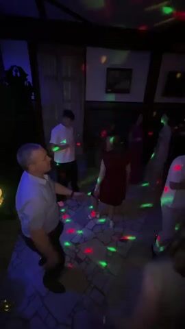 The video shows a group of people dancing in a dimly lit room. The room appears to be a party venue, possibly a banquet hall or a club, with a disco ball casting colorful lights on the walls and floor. The subjects are diverse, including men, women, and teenagers, all engaged in dancing and enjoying themselves. Some individuals are captured in close-up, while others are seen in the background, contributing to the lively atmosphere. The setting is characterized by dark walls and a patterned floor, with some framed pictures or artwork visible on the walls. There are no visible text elements in the video. The time of day is likely evening or night due to the darkness and artificial lighting. The overall mood conveyed is celebratory and joyful, with people expressing themselves through dance. The location context provided suggests this event is taking place in Zavod Mikhailovskii, Russia.