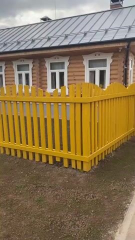 The video pans across a residential property. The house is made of logs and has a grey metal roof with a chimney. A bright yellow wooden fence encloses the front yard. The fence has a gate with a starburst design. The driveway is paved with red and brown bricks. A sign with a nature-themed illustration is visible in the yard. The sky is cloudy.