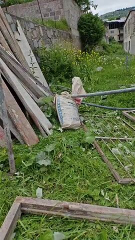 This media file depicts an outdoor scene with various objects and elements of a construction or demolition site. The foreground is dominated by lush green grass and foliage, interspersed with scattered debris. Prominently featured are several pieces of what appear to be discarded wooden planks leaning against each other. To the right of the planks, a large bag, likely containing building materials like cement, is visible. Further into the scene, a rusted metal gate lies on the ground, alongside other metallic structures.

In the background, the scene transitions to a partially constructed or dilapidated building made of stone and concrete. The building has open window frames and doorways, suggesting it is unfinished or abandoned. The surrounding area is also overgrown with grass and wildflowers, indicating that the site may have been left unattended for some time. In the distance, hints of a hillside and other structures can be seen, suggesting a rural or semi-rural setting.

The time of day appears to be daytime, with natural light illuminating the scene. The weather seems overcast, as there are no strong shadows, and the overall lighting is soft. The overall impression is one of disarray and neglect. There is no visible activity of people taking place in the frames provided. The visual cues do not convey any specific emotions, but the setting might evoke feelings of abandonment or the passage of time. No text is clearly legible in the provided frames. The location context provided is Gugark, Armenia.