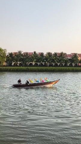 A longtail boat carrying two people is traveling across a river. The boat has colorful decorations mounted along its centerline. The river water is rippled and reflects the sky.  In the background, there's a line of buildings with red tile roofs, separated from the river by a grassy bank and palm trees. The sky is pale blue, possibly indicating early morning or late afternoon light.
