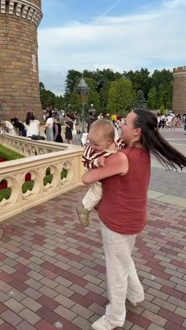 The video shows a woman holding a baby in front of a castle. The woman is wearing a red top and cream-colored pants. The baby is wearing a striped onesie. They are standing on a brick pathway in a plaza. In the background, there is a large castle with turrets and flags, and many people are walking around. The sky is blue with some clouds. There is a decorative railing to the left of the woman. The scene appears to be a tourist attraction, possibly a theme park.