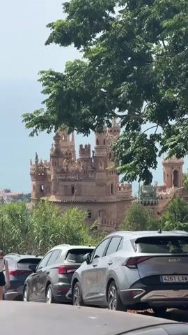 The video shows a view of a castle behind trees and parked cars. In the foreground, the top of a car is visible. Further back, several cars are parked in a row. Two people are walking on the left side of the frame, a man and a woman. The man is wearing a black t-shirt and shorts, and the woman is wearing a white top and shorts. They are walking towards the right of the frame. The castle, made of brick, has multiple towers and is visible in the background. Trees and foliage surround the castle and the parking area. The sky is bright and overcast.