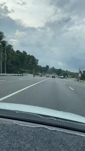 The video, filmed from inside a car, shows a multi-lane highway. The road is grey and has white and yellow painted lines. Vehicles, including a jeepney, truck, and various cars, can be seen traveling in both directions. Lush green trees and foliage line the highway. The sky above is overcast and grey with visible cloud cover. The camera is positioned behind the windshield of the car, and a grey fuzzy dash cover is visible. There is no audio.