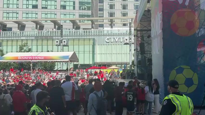 A crowd of people are gathered in an outdoor urban plaza. A large modern building with "CIVIC" visible on it serves as a backdrop. In the foreground, people are sitting and standing, many wearing red clothing and Canada flags, suggesting a sporting event or national celebration. Several people are holding Canadian flags, some unfurling them. To the left, a tent labeled "BUSINESS LOUNGE" is set up. A stage area is visible with people on it, some appearing to interact with the crowd. Security personnel in high-visibility vests are present among the crowd. The weather appears to be sunny.