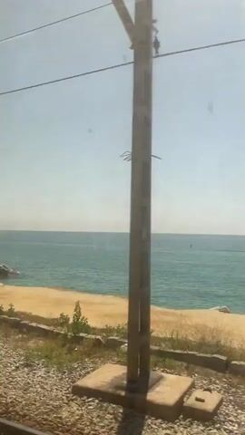 The video captures a view from a moving vehicle, likely a train, looking out at a body of water. The water is a vibrant turquoise color and appears to be a sea or large lake. The sky is a clear, pale blue. In the distance, there are a few small boats or buoys visible on the water. A sandy or light-colored embankment runs along the edge of the water, with sparse vegetation growing on it. In the foreground, there is a gravelly area and more vegetation, suggesting the vehicle is traveling along a track adjacent to the embankment. There are overhead wires visible, reinforcing the idea that this is a train journey. The light suggests it is daytime, likely a clear day. No people are clearly visible.