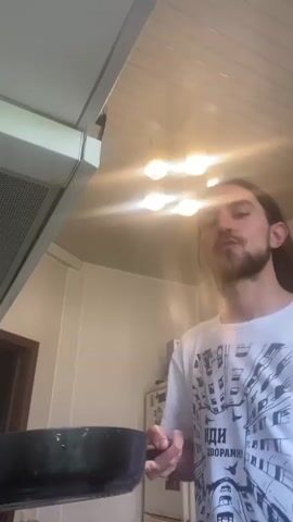 The media file shows a young man with long brown hair and a beard, wearing a white t-shirt with a graphic print. He is in what appears to be a kitchen, indicated by the presence of a range hood, a refrigerator, and ceiling lights. The lighting suggests it is daytime, with sunlight filtering in and the overhead lights on.

The man is holding a frying pan, and in the sequence, he flips something in the pan, suggesting he is cooking. The object being flipped is round and appears to be a pancake or similar food item. He smiles at the camera in the final frames, indicating a playful or satisfied mood. The background shows typical domestic elements, including what looks like a microwave on top of the refrigerator, and a doorway to the left. There is text visible on the man's t-shirt which appears to be in Cyrillic script, with the visible portion reading "ИДИ ДВОРАМИ".