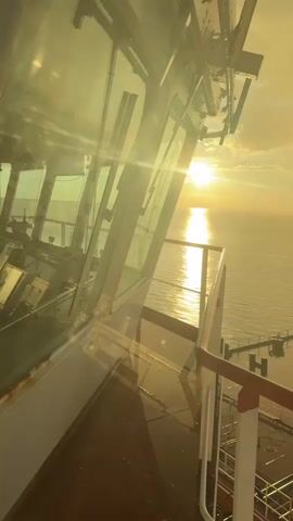 The video shows a view from a ship's bridge during sunrise or sunset. The sun is low on the horizon, casting a golden light across the water and clouds. The bridge itself has large windows, and the reflection of the sun is visible on them. Part of the deck and railing of the ship can be seen in the foreground. The overall scene is serene and picturesque.