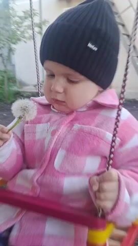 A young child, approximately one year old, sits on a swing outdoors. The child wears a black beanie, a pink and white checkered jacket, patterned leggings, and white sneakers. They hold a dandelion in their hands and bring it towards their mouth. The swing has a red seat and beaded decorations on the ropes. The background shows a building with a window and a metal staircase, along with some greenery and dirt ground.