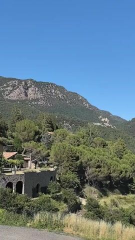 This is a vertical video showing a scenic landscape with mountains and lush greenery. The camera pans from left to right, revealing a wide view of a natural environment under a clear blue sky, suggesting a sunny day with pleasant weather.

In the foreground, a stone structure with arched openings is visible, possibly part of a building or a historical ruin, nestled among trees and vegetation. Further into the scene, rolling hills covered in dense green trees dominate the landscape. The trees appear to be a mix of coniferous and deciduous types, creating a rich texture across the slopes.

As the camera continues to pan, the view expands to encompass more distant mountains, some with rocky outcrops and others cloaked in vegetation. In the mid-ground, a body of turquoise water, likely a lake or reservoir, is visible, adding a splash of color to the otherwise green and brown hues of the natural setting. On the far right, there are signs of human development with what appears to be a building or complex, possibly a hotel or industrial site, situated near the water.

The overall impression is one of tranquility and natural beauty, with the sunlight highlighting the contours of the land and the vibrant colors of the vegetation and water. The time of day appears to be midday, given the bright sunlight and the clear, shadow-free conditions. The location context of Sant Llorenç de Morunys, Spain, suggests a mountainous region known for its natural landscapes. No people or text are visible in the video.
