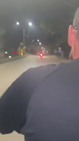 A person wearing a black shirt and glasses rides on a scooter at night. Streetlights line the side of the road. A vehicle with red lights is visible in the distance. The person turns their head briefly to look back at the camera.