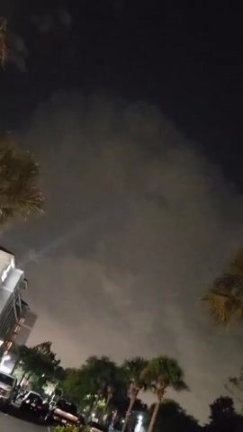 The video shows a nighttime scene with a cloudy sky above. In the lower left portion of the frame, a building with illuminated windows is visible, along with a white van and other vehicles parked nearby. Several palm trees and other trees with green foliage are scattered throughout the lower part of the image. A faint beam of light can be seen in the upper half of the frame, cutting through the clouds. The overall lighting suggests it is dark, with artificial lights from the building and vehicles providing illumination.