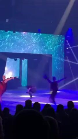A stage performance is taking place with multiple dancers in view, illuminated by purple and white lights. There is a large screen behind them displaying abstract blue and green visuals. The dancers wear various costumes including red dresses and dark outfits. Some are performing acrobatic moves. The audience is visible in the foreground, primarily as silhouettes.