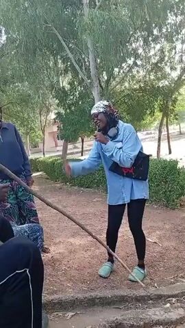 In a park-like setting, a woman in a light blue shirt, black leggings, and light green shoes dances and gestures energetically while holding a microphone to her mouth. She also wears a patterned headscarf and headphones, and has a black bag strapped across her chest. Another woman, wearing a grey jacket and patterned skirt, stands nearby laughing. A person's arm extends a wooden stick towards the woman in blue, while other individuals are seated nearby, partially visible. The background features trees, a building, and a grassy area.