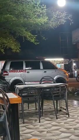 This media file captures a nighttime scene on a street in Keuniree, Indonesia. The focal point is a silver ambulance with red lettering that spells out "AMBULANCE" on its side. The ambulance is parked on the side of the road, with its emergency lights visible on the roof.

In the foreground, there is a table with four black chairs. The table is set with what appears to be a white tablecloth with a red stripe. The ground beneath the table and chairs has a patterned tile floor. To the left of the table, part of another table and chairs are visible, suggesting this is an outdoor seating area, possibly a cafe or restaurant.

Behind the ambulance, there are several buildings and structures, indicating a commercial or urban area. One building has text that is partially visible, reading "GINIA TEMBAKAU", suggesting it might be a tobacco shop or a related business. There are also lights illuminating the scene, typical of a nighttime setting in a populated area.

A person is visible riding a motorcycle to the right of the ambulance, adding a sense of movement and activity to the otherwise still image. The overall mood is calm and ordinary, despite the presence of the ambulance, suggesting it might be parked temporarily rather than actively responding to an emergency. The time of day is clearly night, with a bright light fixture visible overhead, casting a glow on the scene. The leaves of a tree are visible in the upper left corner, suggesting the presence of greenery in the vicinity.