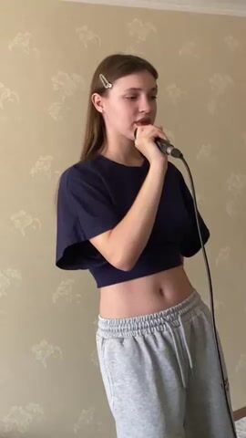A young woman with long, light brown hair and two silver bobby pins is singing into a microphone. She is wearing a dark blue crop top and grey sweatpants. The background is a cream-colored wall with a subtle floral pattern. Her left arm is bent at the elbow and her hand is holding the microphone. She is looking upwards and her mouth is open as if singing.