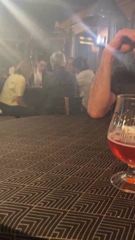 The video shows the interior of a dimly lit bar or restaurant, with a focus on a man at a table and the general atmosphere. The bar has a smoky or hazy ambiance.

In the foreground, a table with a patterned tablecloth holds a half-full glass of reddish liquid, possibly beer or wine. Next to the glass is a black vaping device and a small glass dish. A man in a dark patterned shirt is sitting at the table. He looks toward the right and appears to be talking.

In the background, there are several other tables with people seated, engaged in conversations and interactions. The lighting is low, with light filtering in from unseen sources, creating an atmospheric effect. The interior has wooden elements and a mix of modern and rustic decor.

The overall impression is of a social gathering in a relaxed setting.