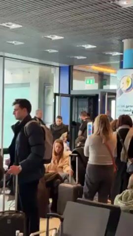 The video depicts an airport scene. Several people are standing or seated with luggage, seemingly waiting. A police officer is standing near an open doorway, observing the crowd. He is wearing a black uniform with "POLICE" printed on the back. An exit sign is visible above the doorway. The environment appears to be a terminal or waiting area.