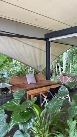 The video shows an outdoor seating area with a lush green background. The seating area appears to be part of a cafe or restaurant with wooden furniture, including tables, benches, and chairs. The floor is dark-colored wood, and a stream or river flows in the background amidst the trees. The camera pans from an area with a couch and plants towards a more open area with additional tables and benches. Two individuals are briefly visible, one adjusting their posture on a chair. The atmosphere seems serene and rainy, with the sound of flowing water.