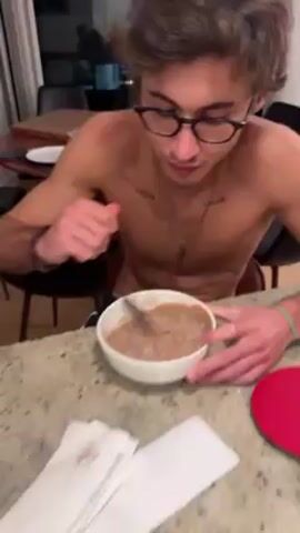 A shirtless young man wearing glasses is eating from a white bowl with a spoon while standing at a countertop. The bowl contains a brown mixture. He is looking away from the bowl while chewing. A white napkin is on the counter next to the bowl. The countertop is speckled and appears to be made of granite. In the background are dining chairs and part of a dining table. The lighting appears to be indoor lighting. The man has tattoos on his chest and is wearing a necklace.
