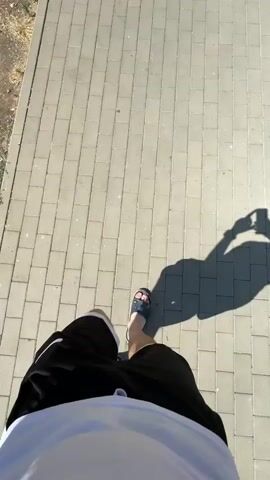 A first-person perspective video shows a person walking on a paved sidewalk. The person is wearing black shorts with white trim and a white t-shirt. Their left foot, wearing a sandal with colorful circular designs, is in focus, while their right foot is out of frame. A long shadow of the person, holding what appears to be a phone or camera, stretches across the sidewalk. The environment is outdoors, with sunlight casting a strong shadow. The sidewalk is made of rectangular gray pavers. To the left of the sidewalk, there is dry, sparse vegetation. The camera angle is looking down at the person's legs and feet as they walk away from the camera. The video captures the movement of the person's legs and the changing position of their shadow over the course of a few seconds.