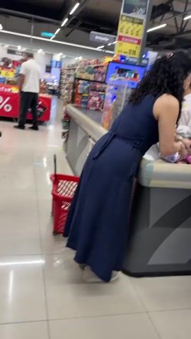 A woman in a dark blue dress stands at a supermarket checkout counter, leaning over to interact with a cashier. A red shopping basket sits on the floor next to her. In the background, other shoppers and cashiers are visible. The environment appears to be a brightly lit supermarket aisle with shelves stocked with products.
