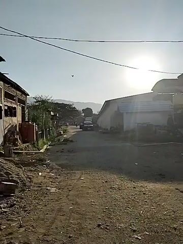 The video shows a dusty, unpaved road with sparse vegetation and buildings on either side. The sun is shining brightly in the sky, casting long shadows. In the foreground, the road is covered in gravel and dirt. There are some objects scattered on the ground, possibly debris. Further down the road, several vehicles are visible, including a dark-colored pickup truck, a motorcycle, and possibly another car. The buildings appear to be simple structures, some with corrugated metal roofs. The overall impression is of a rural or semi-developed area. The camera is moving, suggesting it is attached to a vehicle or being held by someone walking. The video is low resolution and grainy.