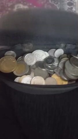 A close-up, blurry video shows a black pouch filled with assorted coins. The pouch appears to be made of fabric, possibly a coin purse or a small bag. The coins are a mix of different sizes, colors, and apparent materials, suggesting they are of various denominations and possibly from different countries. Some coins are a light silver color, while others are a brass or gold hue. A person's fingers, with manicured nails, briefly enter the frame from the left side of the pouch, reaching for the coins. The lighting is dim, and the focus shifts throughout the video, making it difficult to discern specific details on the coins or the contents of the pouch. The background is dark and out of focus.