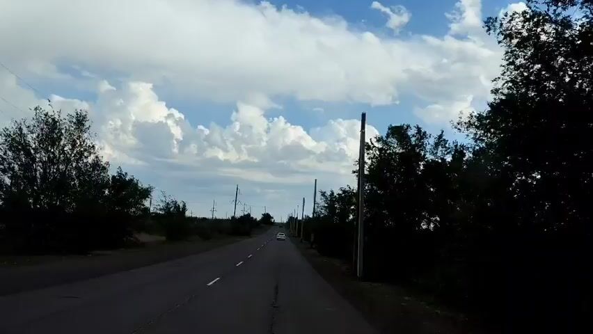 A car drives down a paved road lined with trees and utility poles under a sky filled with white clouds.
