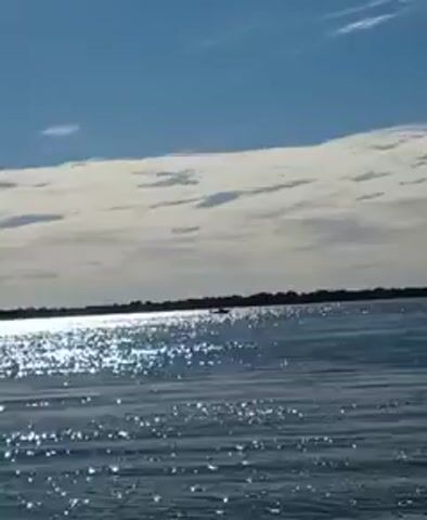 The video shows a distant view of water with sunlight glinting off the surface. A boat is visible in the distance, appearing as a dark silhouette against the water and the horizon of land. The sky is blue with a layer of white clouds. The camera seems relatively stable.