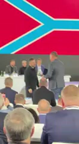 The video captures an indoor event taking place in Barvinok, Ukraine. In the foreground, numerous individuals are seated, observed from behind, with their attention directed towards the front. Behind them, a panel of individuals is seated at a white table, some of whom are actively applauding.

The central figure, Ukrainian President Volodymyr Zelenskyy, dressed in a black long-sleeved shirt, stands facing the audience. He is seen shaking hands with another man before a woman approaches him to present a blue folder or plaque. Zelenskyy accepts the item, smiles, and gestures towards the applauding audience, acknowledging their warm reception. The atmosphere appears positive and celebratory, suggesting an official ceremony or recognition event.

Behind the panel and Zelenskyy, a large screen is prominently displayed. Initially, it features a red background with a distinct light blue, inverted 'Y' shape, reminiscent of a stylized flag. The screen's display then transitions to a solid bright green color. No specific text is clearly discernible on the screen or the presented item. The event appears to be well-attended, with the audience demonstrating approval through applause.
