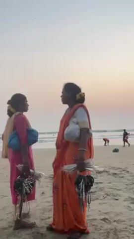 Two women stand on a sandy beach at sunset. The woman on the left wears a pink outfit with a tan scarf. The woman on the right wears an orange sari with a white blouse. Both women hold bunches of necklaces in their hands. They seem to be speaking to one another, then the woman in the sari looks at the camera. In the background, there are people on the beach and in the water, and the sky has hues of pink and orange from the sunset.