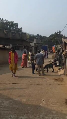 The video shows a street scene in a village or small town. Several people are walking along a paved street. A child stands next to a man, who is holding his hand.  A goat is nearby. Other adults stand or walk close by. The buildings are simple, some appearing to be in disrepair. Trees and a hill are visible in the background. The sky is overcast.