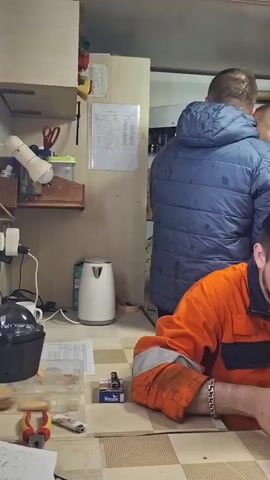 A group of men are in a small break room. One man in an orange jumpsuit sits at a table, looking at his phone. There is a cup and an ashtray on the table in front of him. Another man in a blue jacket stands behind him. To the right of the man with the phone, an older man in a dark blue jumpsuit sits at the table, talking. To his right and further back, a man in a red jumpsuit sits on a chair against the wall. There are several pieces of work gear hanging on a hook on the wall. In the background, there is a poster that reads "First Aid."