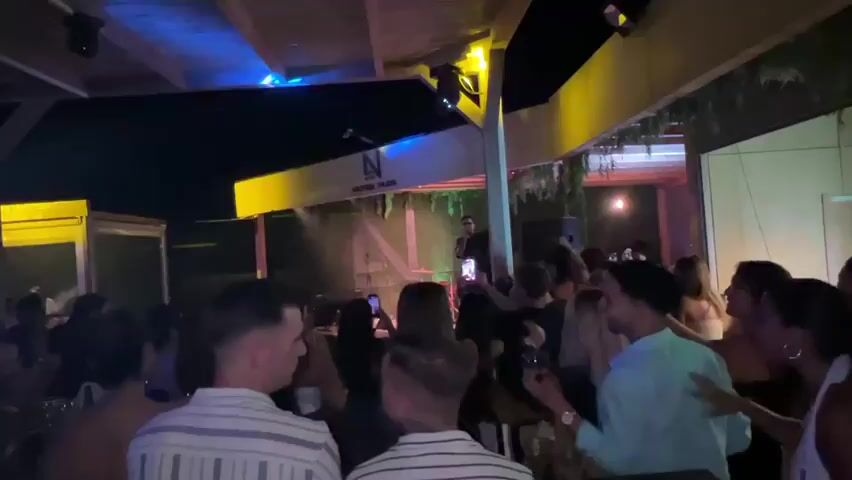 A nighttime outdoor event or party is depicted. A crowd of people are gathered, many facing a stage area. On the stage, a person is standing and appears to be speaking into a microphone. Stage lights are visible, including yellow and purple lighting, and there is smoke or fog in the air around the stage. The environment suggests an entertainment venue, possibly a club or outdoor bar, with a covered roof structure supported by wooden beams and greenery. The attendees are dressed in casual and semi-formal attire, with some holding drinks. The overall atmosphere is lively, with people interacting and some dancing.