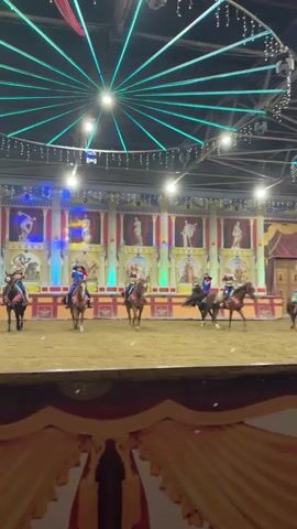 Five people, dressed in costumes that resemble ancient Greek or Roman attire, are riding horses in a circular arena. The arena has a dirt floor and is surrounded by a stage with colorful backdrops depicting classical architecture and statues. Above the arena, there is a ceiling with green lights arranged in a starburst pattern and some hanging disco balls. The people on horseback appear to be performers, and they are positioned at various points around the arena. The lighting is dim, with spotlights illuminating the performers and the stage area. The overall atmosphere suggests a live performance or show.