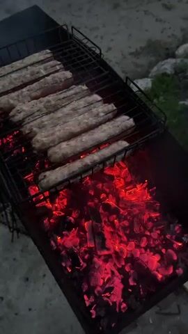 A close-up shot shows several raw sausages grilling over glowing red coals in a barbecue grill. The sausages are laid out in rows on a black metal grate, and the coals beneath them are intensely hot, emitting a vibrant red glow. The background is slightly blurred, suggesting an outdoor setting with sand or dirt on the ground and some greenery in the distance. The lighting is dim, indicating it might be dusk or nighttime. The camera angle is from above, looking down at the grill.