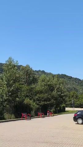The video shows a parking lot on a sunny day. A black car is parked on the right side of the frame. A young man in a white t-shirt and dark pants is standing next to a white dispenser with a red stripe, giving a thumbs down. In the background, there are trees and a hill. There are also red benches in the foreground.