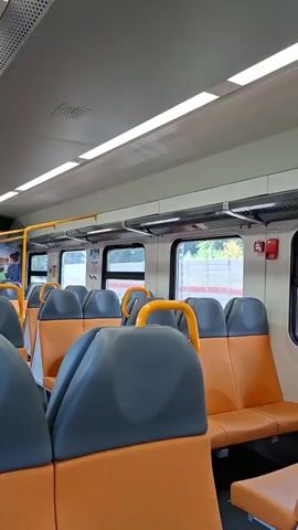 The video shows the interior of a train carriage with rows of empty seats. The seats are upholstered in a dark grey material on the backrests and a bright orange material on the seat cushions. Each seat has a yellow handle attached to its back. The carriage is well-lit by fluorescent lights running along the ceiling. Large windows line the side of the carriage, offering a view of trees and greenery outside. Various safety features, such as a red fire alarm box, are visible on the walls. The perspective of the camera suggests someone is filming while walking through the carriage, or possibly while seated and looking around. The overall impression is of a clean and modern train interior.