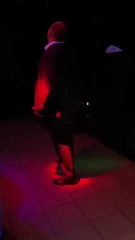A man in a dark suit and white shirt is dancing in a dark room with colored lights. A woman in a pink outfit walks past in the foreground.