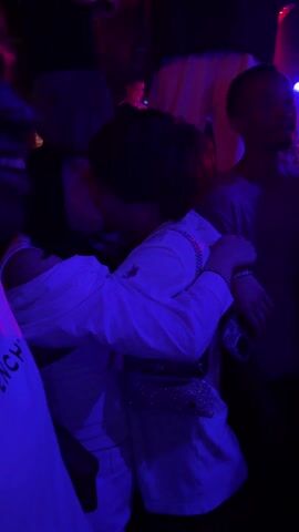 The video shows two people embracing and appearing to kiss in a crowded club or party environment. They are surrounded by other people, but the focus is on them. The lighting is dark and mainly blue, which makes it difficult to see details. The person on the left is wearing a white top with an off-the-shoulder design. The person on the right is wearing a white shirt with a designer logo on the upper chest. They have a small purse or fanny pack on. This person also has a chain and bracelets on. The background consists of other individuals and the general ambiance of a crowded place with moving lights.