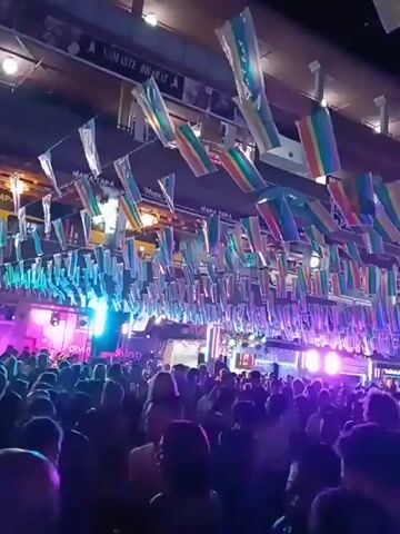 The video shows a large crowd of people gathered in an outdoor space at night. The scene is decorated with many rainbow flags hanging overhead. Bright green and blue lights illuminate the crowd and a stage area. The people are moving and appear to be enjoying a party or celebration. The overall atmosphere is festive and energetic.