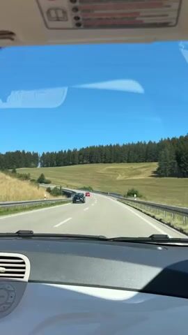 The video begins from inside a car, looking out the windshield on a sunny day. The perspective is from the driver's seat, showing the dashboard and the top edge of the windshield. A black car is visible on the road ahead, slightly to the left, and a small red car is further ahead in the right lane. The road curves to the right, with a barrier on the right side and a lush green landscape of trees and rolling hills on either side of the road. The sky is a clear, bright blue. As the video progresses, the perspective shifts slightly, and the red car is now more prominent in the center of the frame as the car filming it continues down the road. The landscape remains consistent, with the road continuing to curve through the scenic countryside. The video ends as the car continues on its journey.