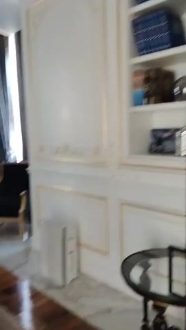 The video pans across a room with ornate white paneled walls featuring gold trim. On the left, a bookshelf is built into the wall, filled with blue-bound books and decorative objects. To the right, heavy grey curtains cover a large window, with a glimpse of a chandelier visible on the ceiling above. In the foreground, a dark upholstered armchair and a small round table with a lamp are visible. The room appears to be a study or library, decorated in a classical style.
