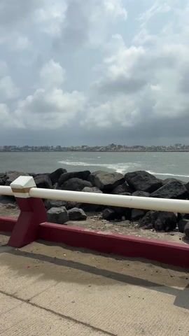 The video shows a view of a harbor from behind a red and white barrier. Large dark rocks line the edge of the harbor, and in the distance, a cityscape is visible under a cloudy sky. The sea is choppy with whitecaps visible on the waves. The camera is static and there are no people in the frame.