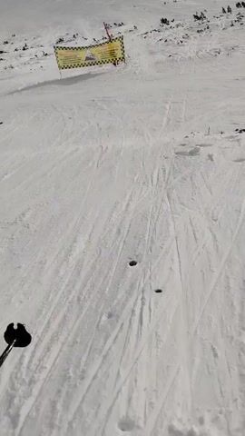 The video shows a first-person perspective skiing on a snowy slope. The camera moves over the snow towards a yellow and black checkered banner with the words "WARNING DANGER OF AVALANCHES! PLEASE USE ONLY THE GROOMED AND MARKED SKI RUNS. IN CASE OF AN INCIDENT THE RESPONSIBILITY IS YOURS" written on it. Beyond the banner, the snow covered slope continues towards more mountains.