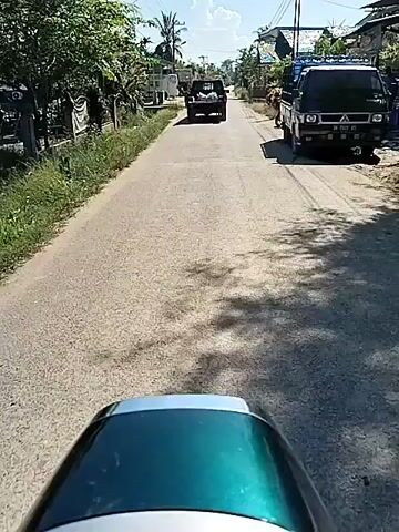 The video shows a first-person perspective from a moving vehicle, likely a scooter or motorcycle, traveling down a paved road. The road is lined with houses and vegetation on either side, typical of a residential area in a tropical or subtropical location. A black pickup truck is driving ahead, with what appears to be a load in the truck bed. The sky is bright and clear, indicating a sunny day.