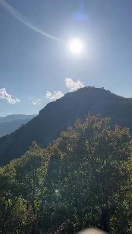 The video shows a panoramic view of a mountainous landscape on a clear, sunny day. The sun is high in the sky, casting a bright light and creating lens flares. In the foreground, lush green trees and bushes with some autumnal foliage are visible. Behind them, a steep, rocky mountain slope rises, partially in shadow. Further in the distance, rolling hills and mountains recede into the haze, with a river or lake visible in a valley. The sky is a vibrant blue with a few small, white clouds. The camera pans slowly from right to left, revealing more of the expansive mountain range and the water body. There are no people or animals visible in the scene.