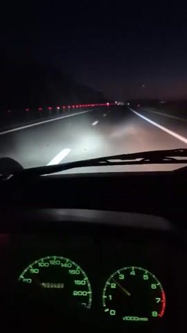 The video is shot from the perspective of a driver at night. The windshield of a car is visible, along with the dashboard featuring illuminated gauges. The speedometer shows a speed around 100, and the tachometer shows the engine running at approximately 2000 RPM. The headlights are on, illuminating the road ahead. Red lights from other vehicles are visible in the distance, suggesting other cars on the highway. The scene depicts driving at night on a road with clear lane markings and a guardrail visible on the left side.
